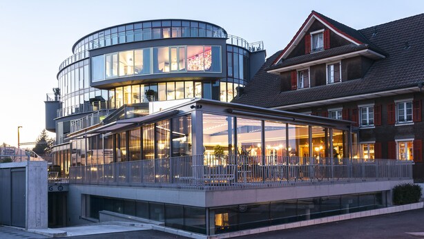 A modern building with a curved glass facade, a covered outdoor seating area, and a traditional building with red shutters in the background. - Hirschen Oberkirch  (Oberkirch)