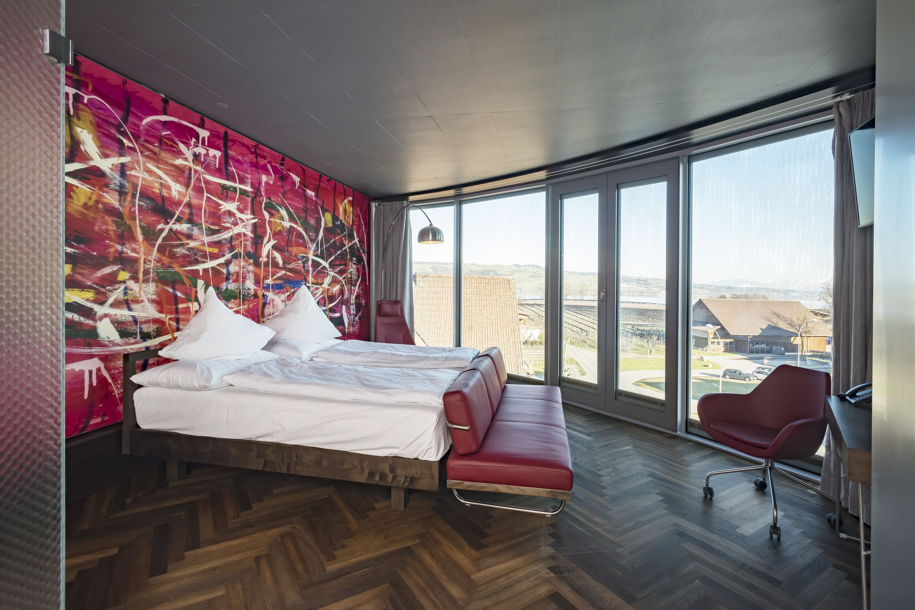 Ett modernt hotellrum med en stor säng, en röd fåtölj och ett stort fönster med utsikt över staden.