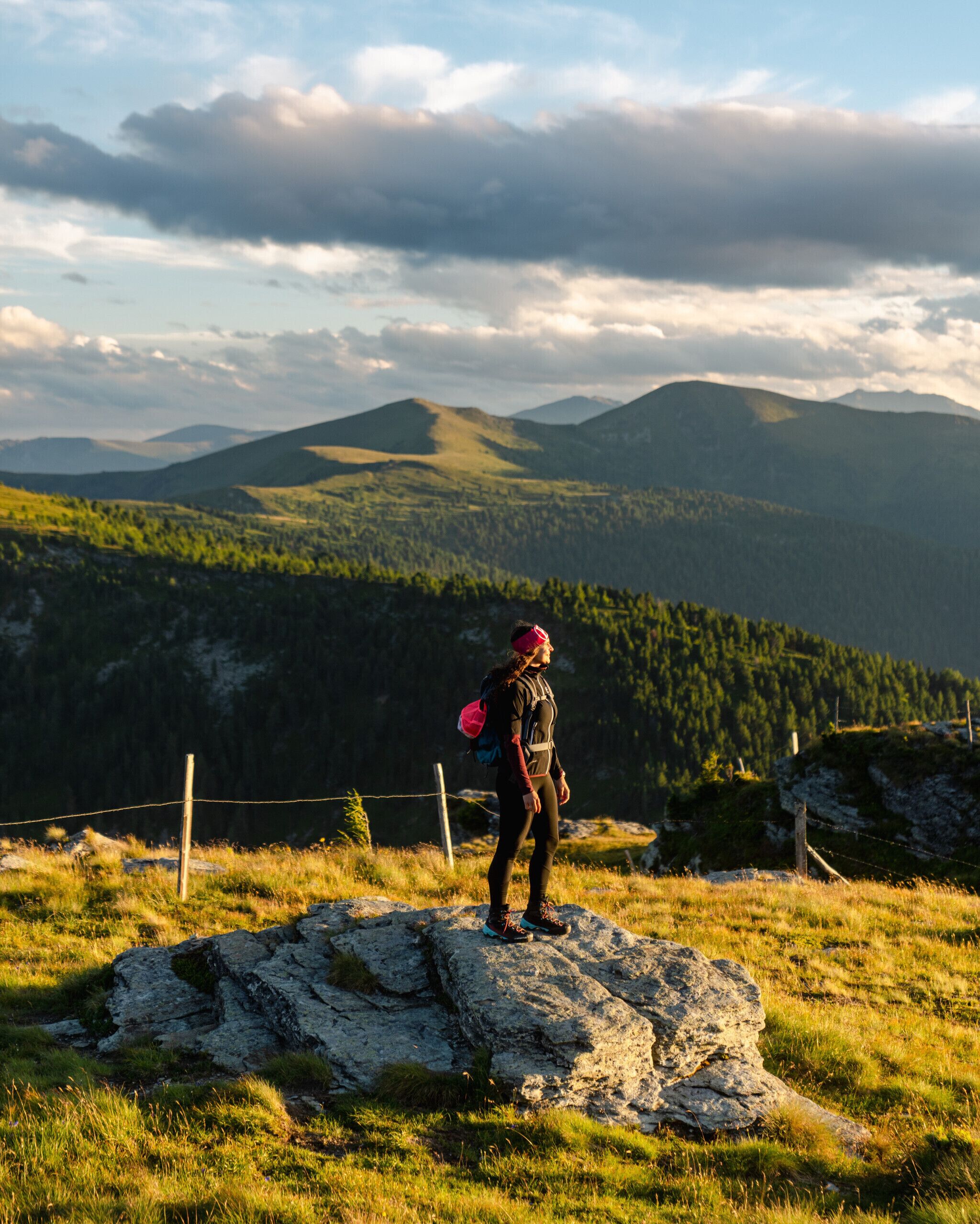Eine Person steht mit Rucksack auf einem Felsen und blickt auf eine gebirgige Landschaft. Im Vordergrund ist ein Zaun zu sehen.