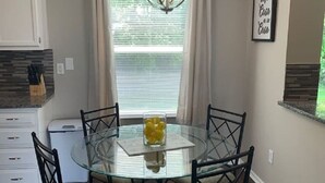 Un comedor con mesa de vidrio rodeada de sillas, una lámpara de araña, una ventana con cortinas y una zona de cocina con armarios y encimera.