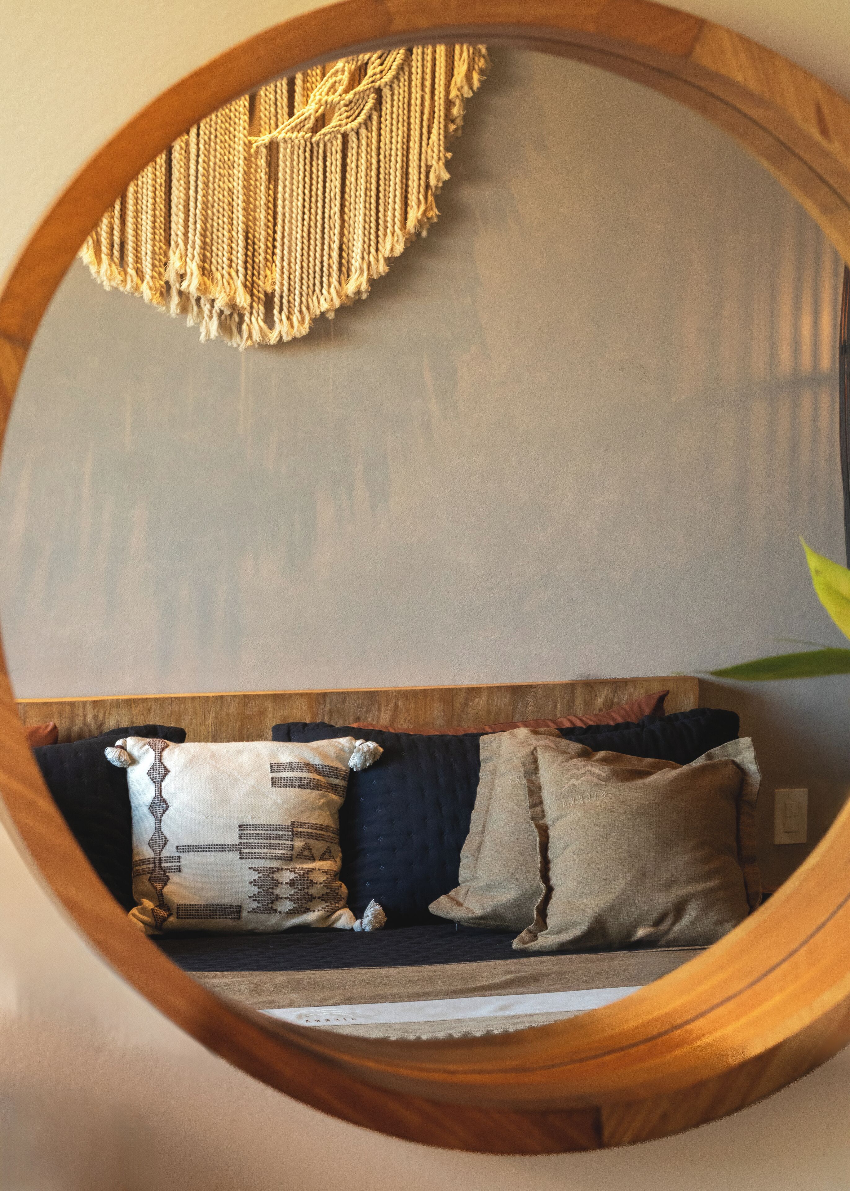 Un espejo redondo con marco de madera que refleja una habitación con un sofá azul oscuro, almohadas estampadas y un tapiz de macramé en la pared.