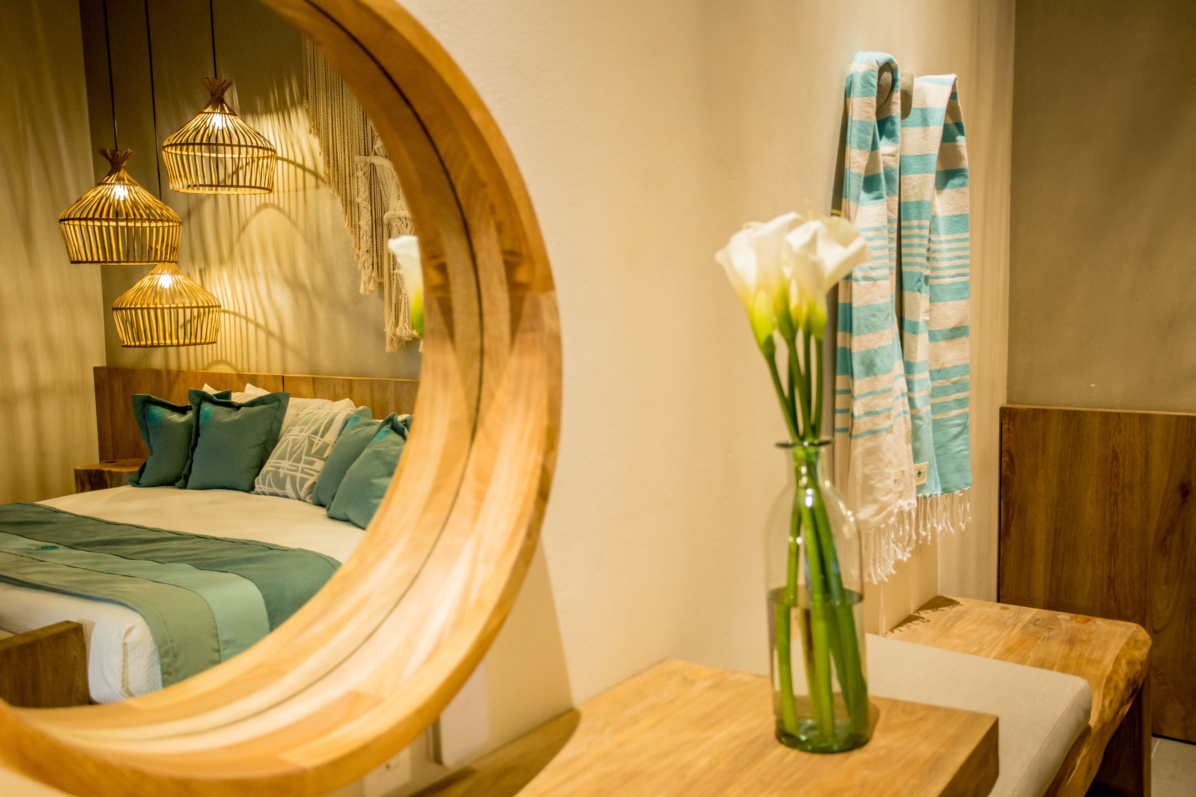 Un dormitorio con cabecera de madera, cama con ropa de cama verde, mesita de noche de madera, espejo, florero con flores blancas y una toalla rayada en azul y blanco.