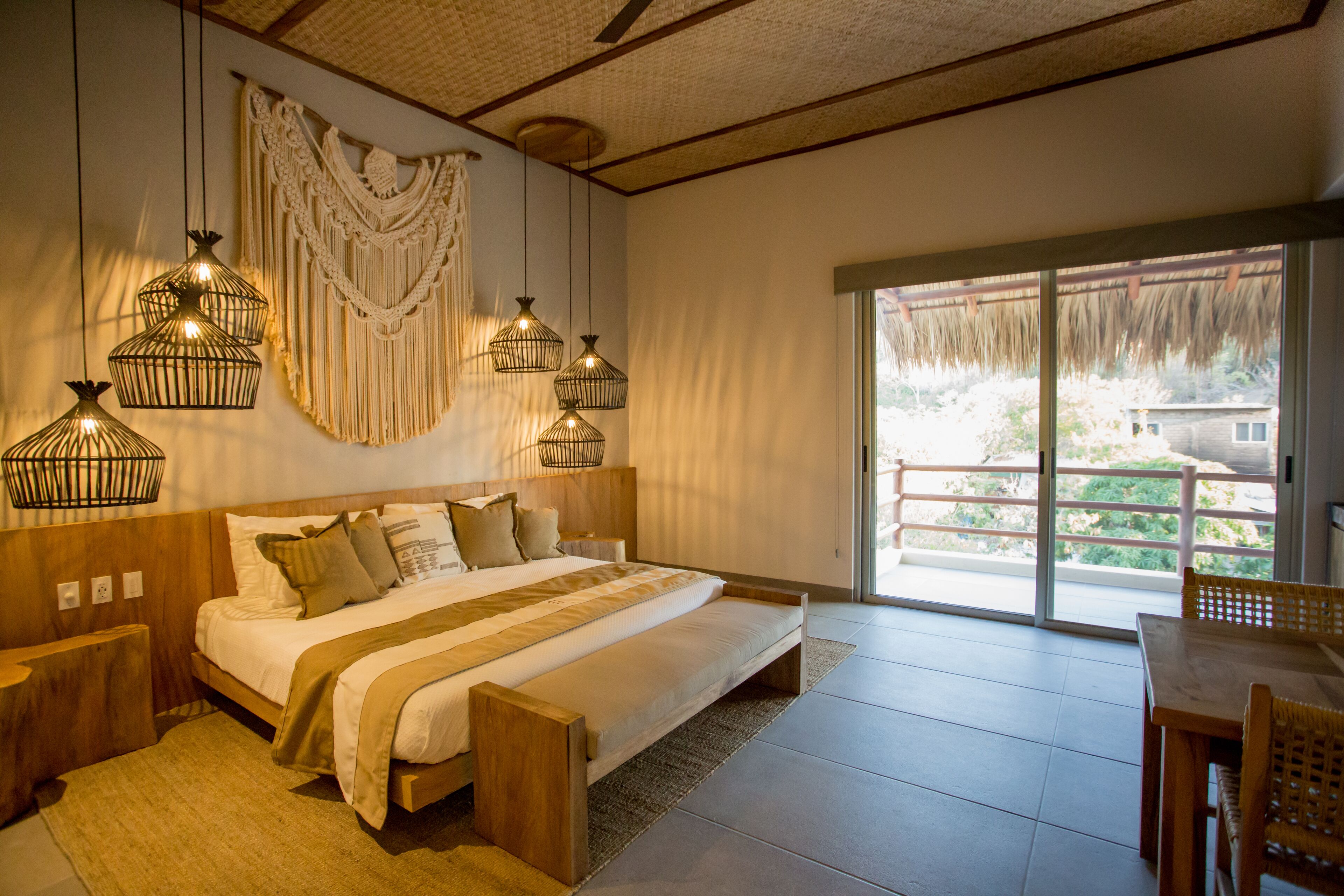 A bedroom with a large bed, wooden headboard, hanging light fixtures, and a view through a sliding glass door to an outdoor area.
