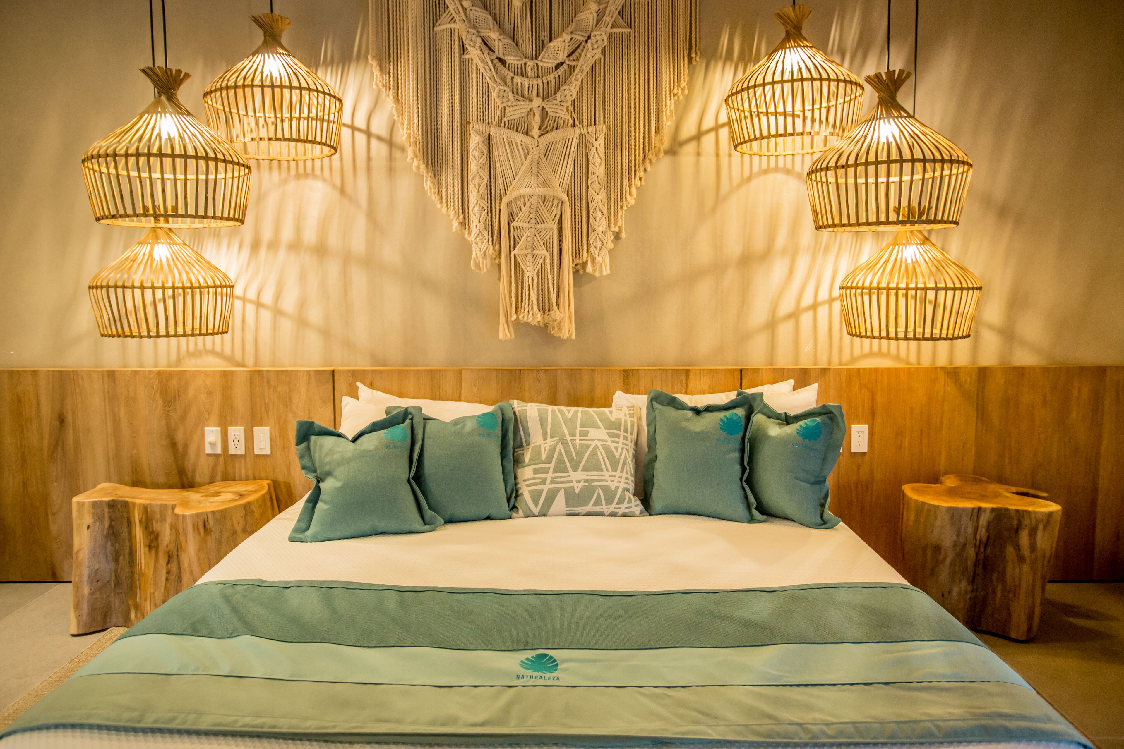Una cama con colcha verde, varios almohadones y un tapiz de macramé en la pared.
