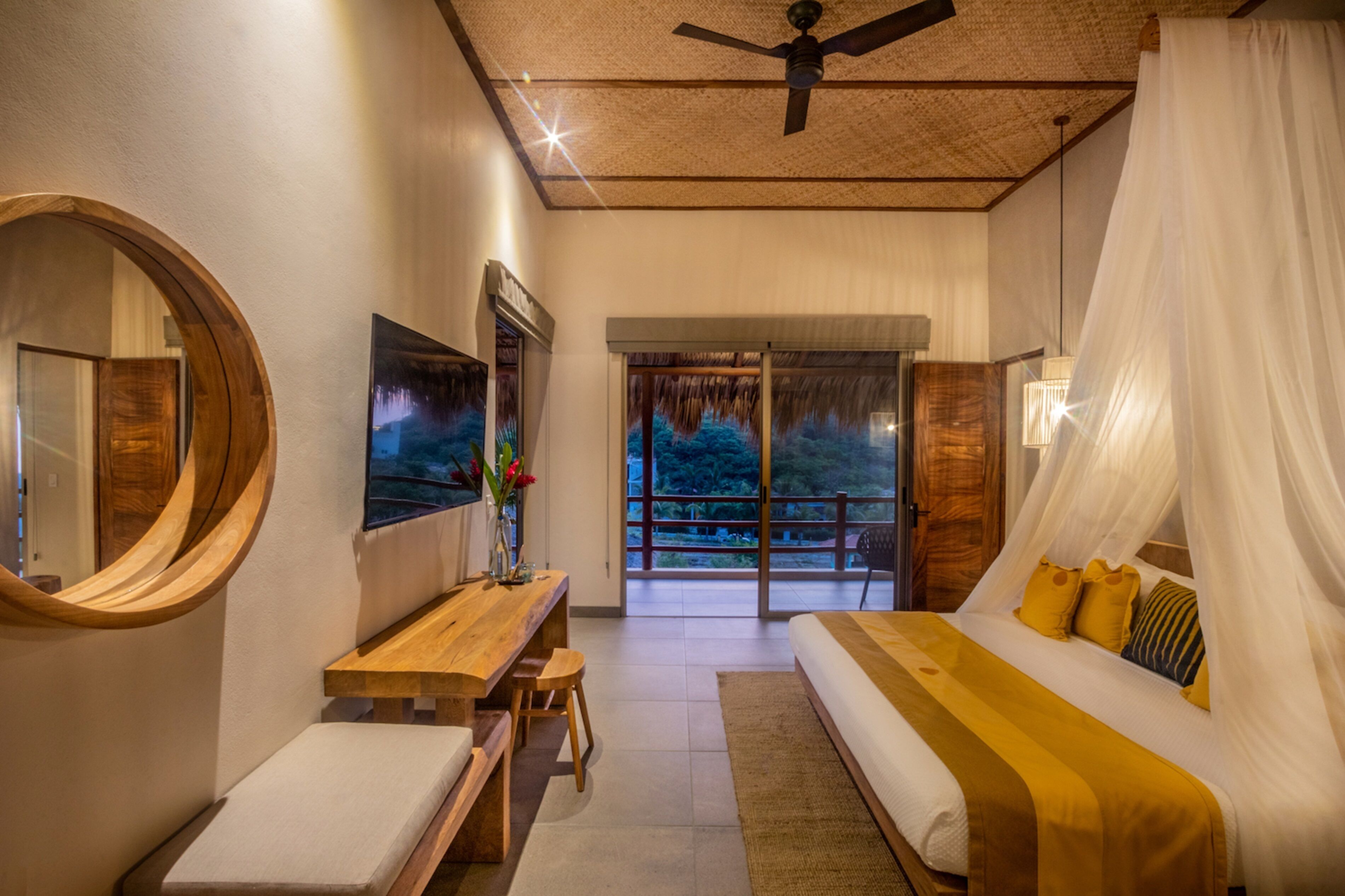 A bedroom with a bed, a desk, a chair, a mirror, and a view of the outdoors.