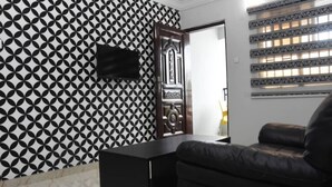 Una habitación con pared estampada, sofá negro, puerta de madera y televisor montado en la pared.