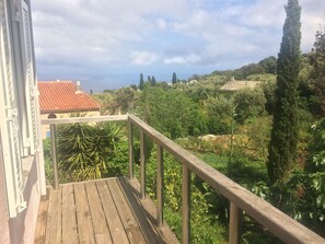 A wooden deck with a metal railing overlooking a lush garden with various plants and trees, and a building with a red-tiled roof in the background.
