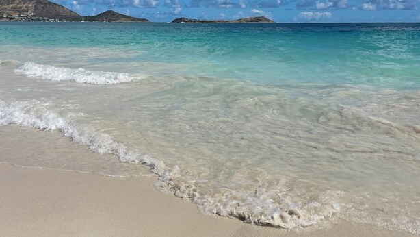 A beach with clear turquoise water, gentle waves, and distant hills. - Beachfront Orient Bay 1‑BR Condo • Mont Vernon • Balcony • Pool • Fast Wi‑Fi (Cul de Sac)