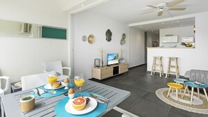 A modern, well-lit living room with a dining area, featuring a kitchen in the background, a ceiling fan, and a TV on a wooden cabinet. - Beachfront Orient Bay 1‑BR Condo • Mont Vernon • Balcony • Pool • Fast Wi‑Fi (Cul de Sac)