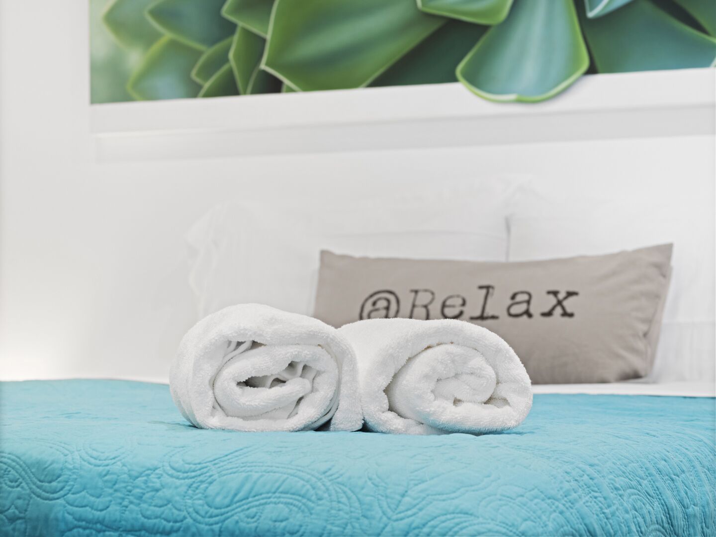 A neatly made bed with a blue quilt, a rolled white towel, and a pillow with "@Relax" text.