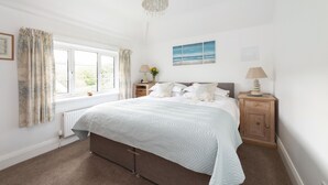 A bedroom with a large bed, two bedside tables, a window with curtains, a radiator, and a chandelier. - Apple Blossom - Chideock, Bridport (Chideock)