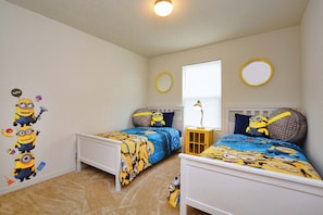 A bedroom with two beds, each with a blue and yellow bedspread featuring minion characters. There are two round mirrors on the walls, a window with blinds, and a small table with a lamp.