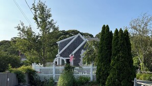 Une maison avec une clôture en bois peinte en blanc, une allée en gravier et un petit jardin où poussent différentes espèces de plantes, ainsi qu’un grand conifère.