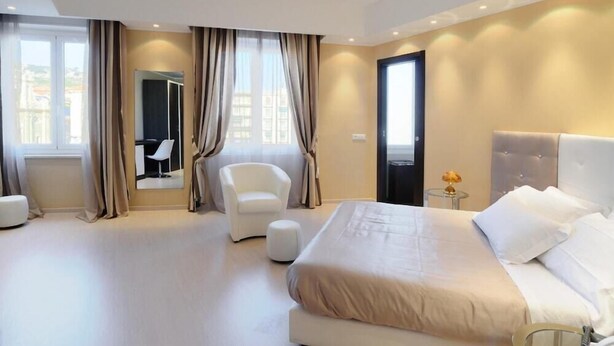Ein modernes Hotelzimmer mit einem großen Bett, einer Sitzecke mit Sessel und Hocker, einem kleinen Tisch mit Lampe und einem großen Fenster mit Vorhängen.