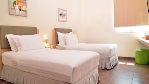A hotel room with two beds, a window with blinds, a green trash bin, and artwork on the walls.