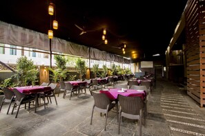 An outdoor restaurant with tables covered in purple tablecloths, wicker chairs, and hanging lantern-style lights. - Fabhotel Arya Regency Hinjewadi (Paud)
