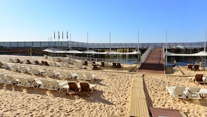 En strand med rækker af solsenge og parasoller, en træbro og et overdækket område med udsigt over vandet.