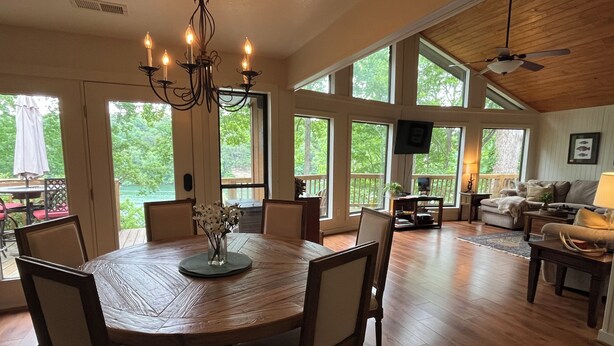 A spacious living area with a wooden dining table, comfortable seating, large windows, and a ceiling fan.