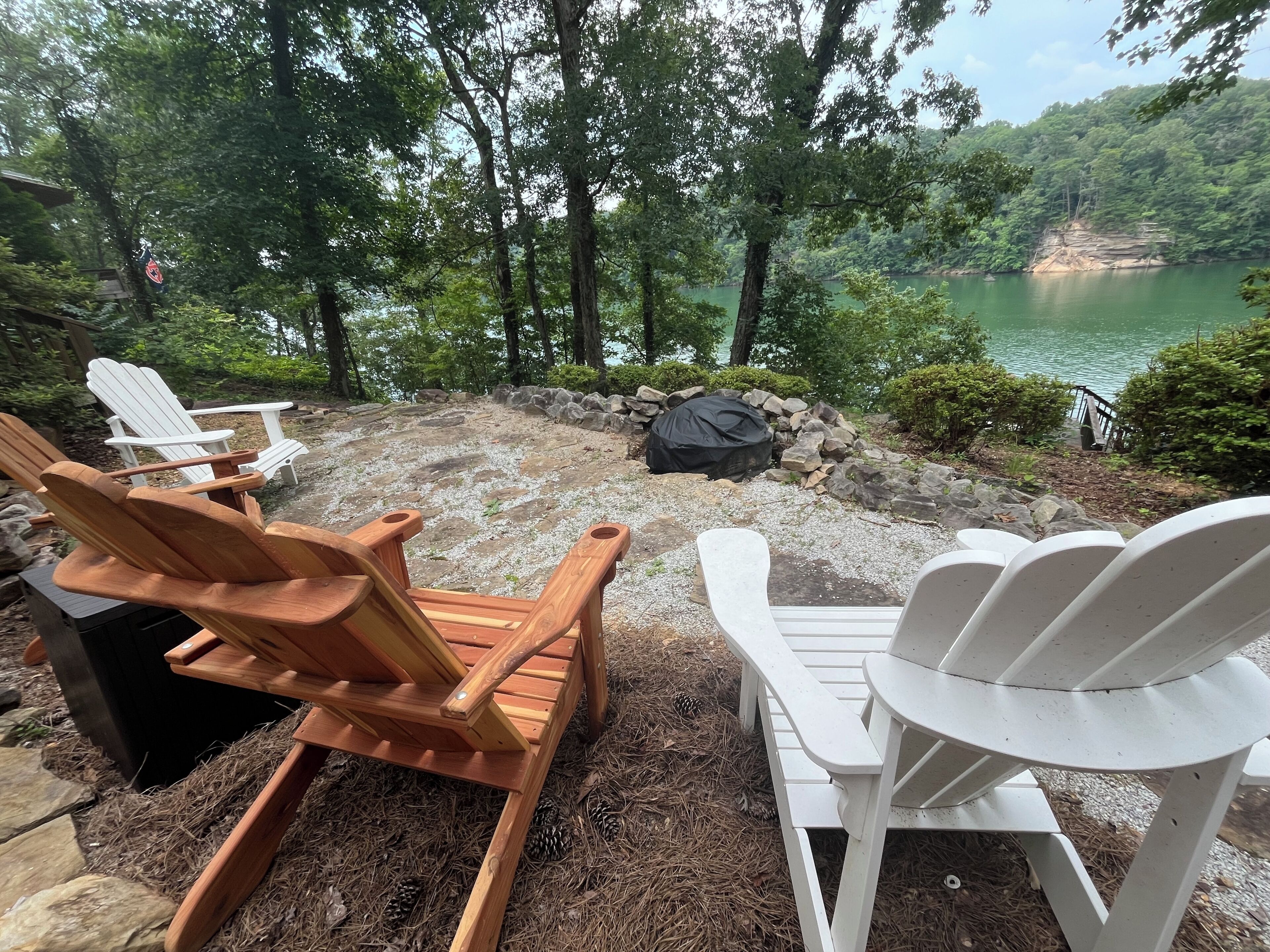 Outdoor seating with Adirondack chairs, a stone fire pit, and a view of a lake surrounded by trees.