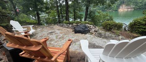 Outdoor seating with Adirondack chairs, a stone fire pit, and a view of a lake surrounded by trees.