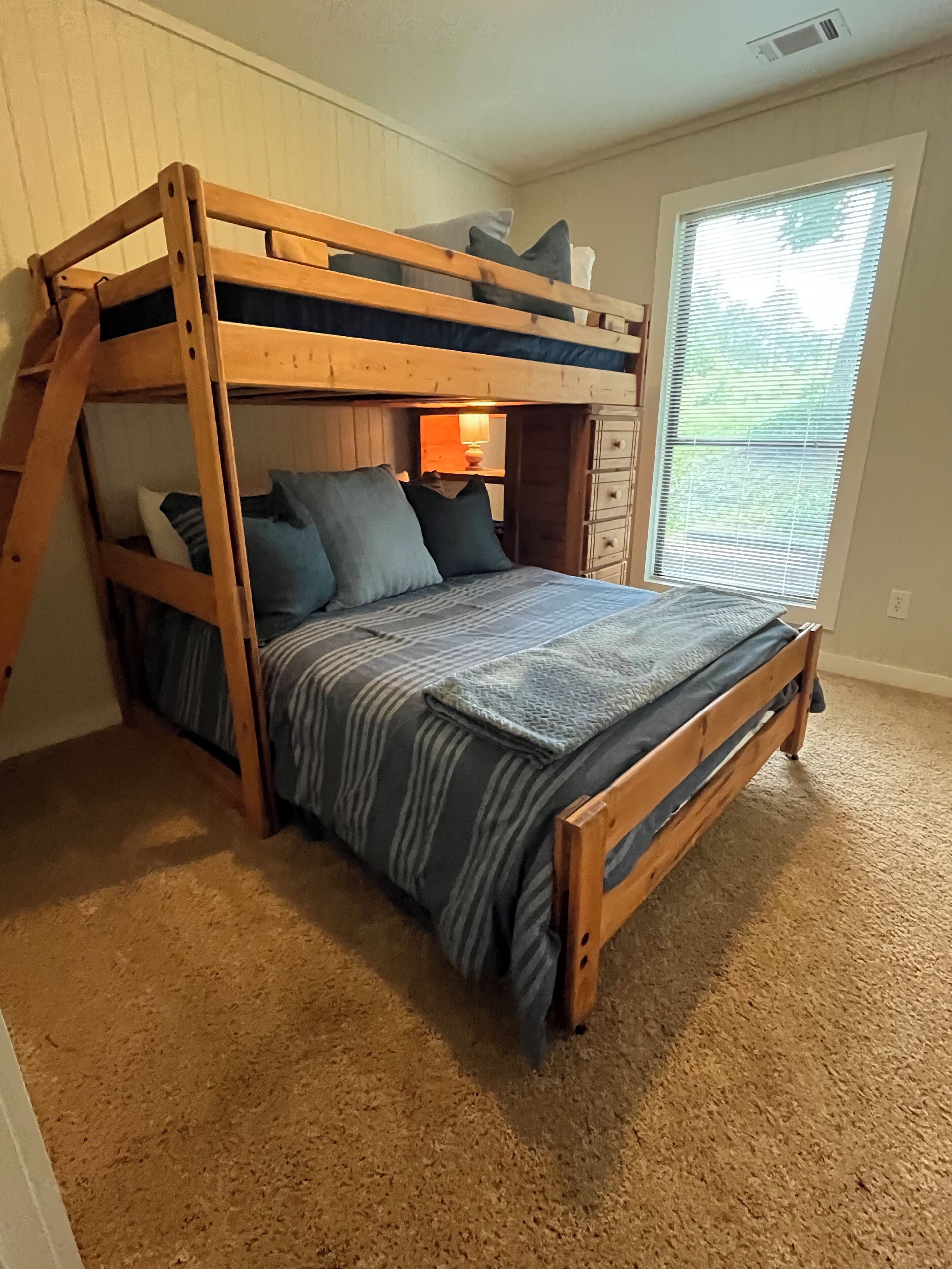 A bunk bed with a wooden frame, a striped bedspread, and pillows. There is a window with blinds and a lamp on the nightstand.