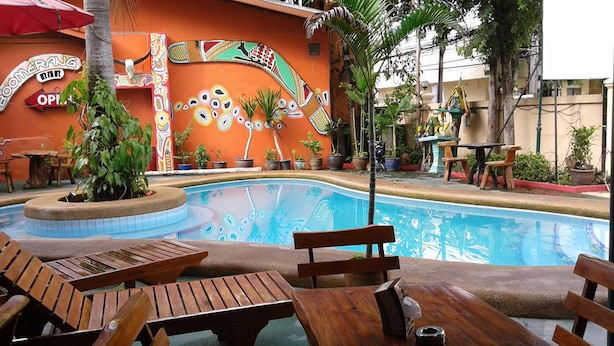 Zona de piscina con mesas y sillas de madera, una pared decorativa con diseños, y un letrero que dice 'Boomerang Bar'.