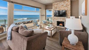 A spacious living room with a stone fireplace, a flat-screen TV, and a view of the ocean. - Guest Favorite! Fire Ring on Sand Walk to Village (Oceanside)