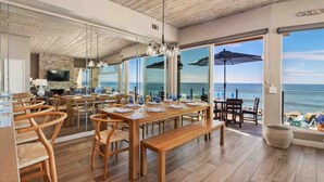 A spacious dining area with wooden tables and chairs, large windows offering a view of the ocean, and a modern interior design. - Guest Favorite! Fire Ring on Sand Walk to Village (Oceanside)