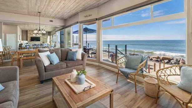 A spacious living area with a large window overlooking the beach, a cozy seating arrangement, and a dining area with wooden furniture. - Guest Favorite! Fire Ring on Sand Walk to Village (Oceanside)