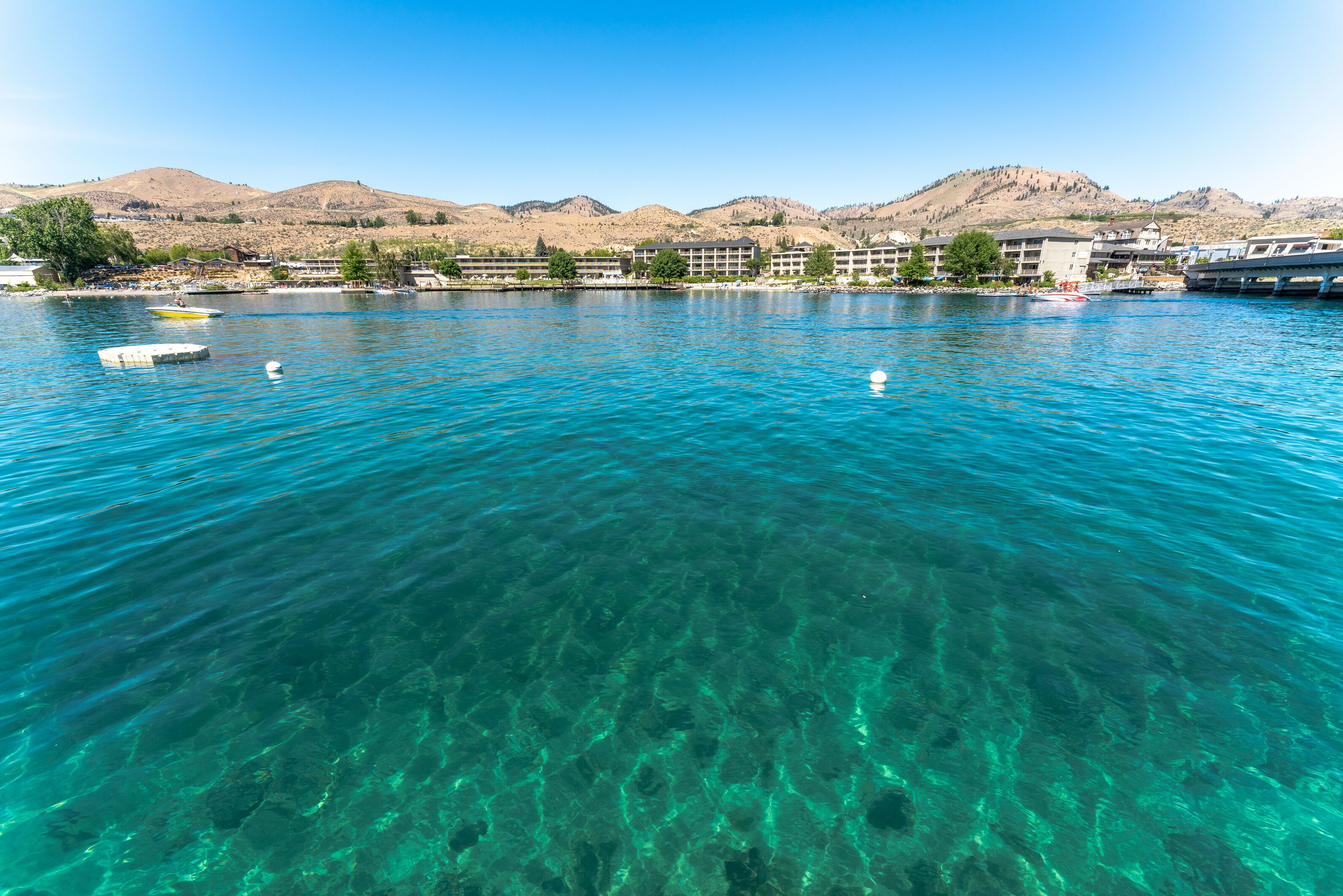 A clear lake with boats, buildings, and mountains in the background.