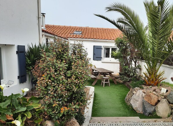 A garden with a wooden table and chairs, a white house with a tiled roof, and various plants.