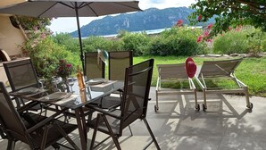 An outdoor dining area with a glass table and chairs, a wine bottle, and a view of mountains and water. - Villa Tout Confort 400 m de la Plage (Porto-Vecchio)