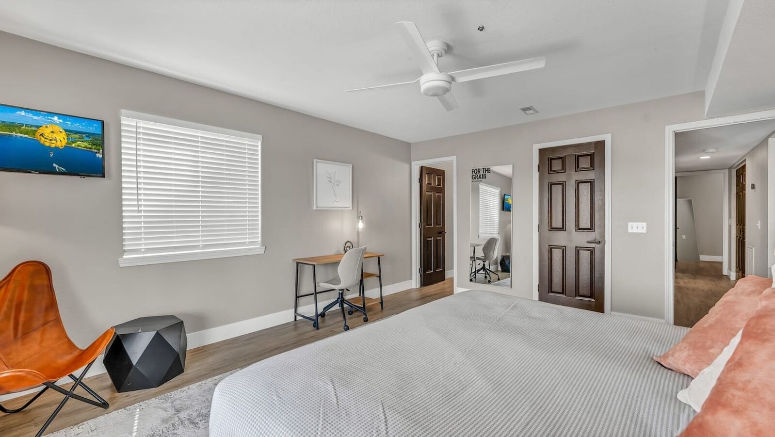 A bedroom with a large bed, a desk with a chair, a ceiling fan, and a TV on the wall.