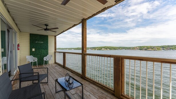 Varanda coberta ao ar livre com vista para um lago, com mesa e cadeiras, ventoinha no teto e parede verde.