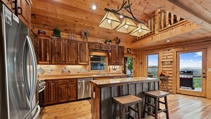 A rustic cabin-style kitchen with wooden cabinets, a stainless steel refrigerator, a large island with stools, and a view of the outdoors through a glass door. - Cherokee Vista - Pet Friendly | Mountain Views | Hot Tub | Shuffleboard | Fire Pit (Blue Ridge)