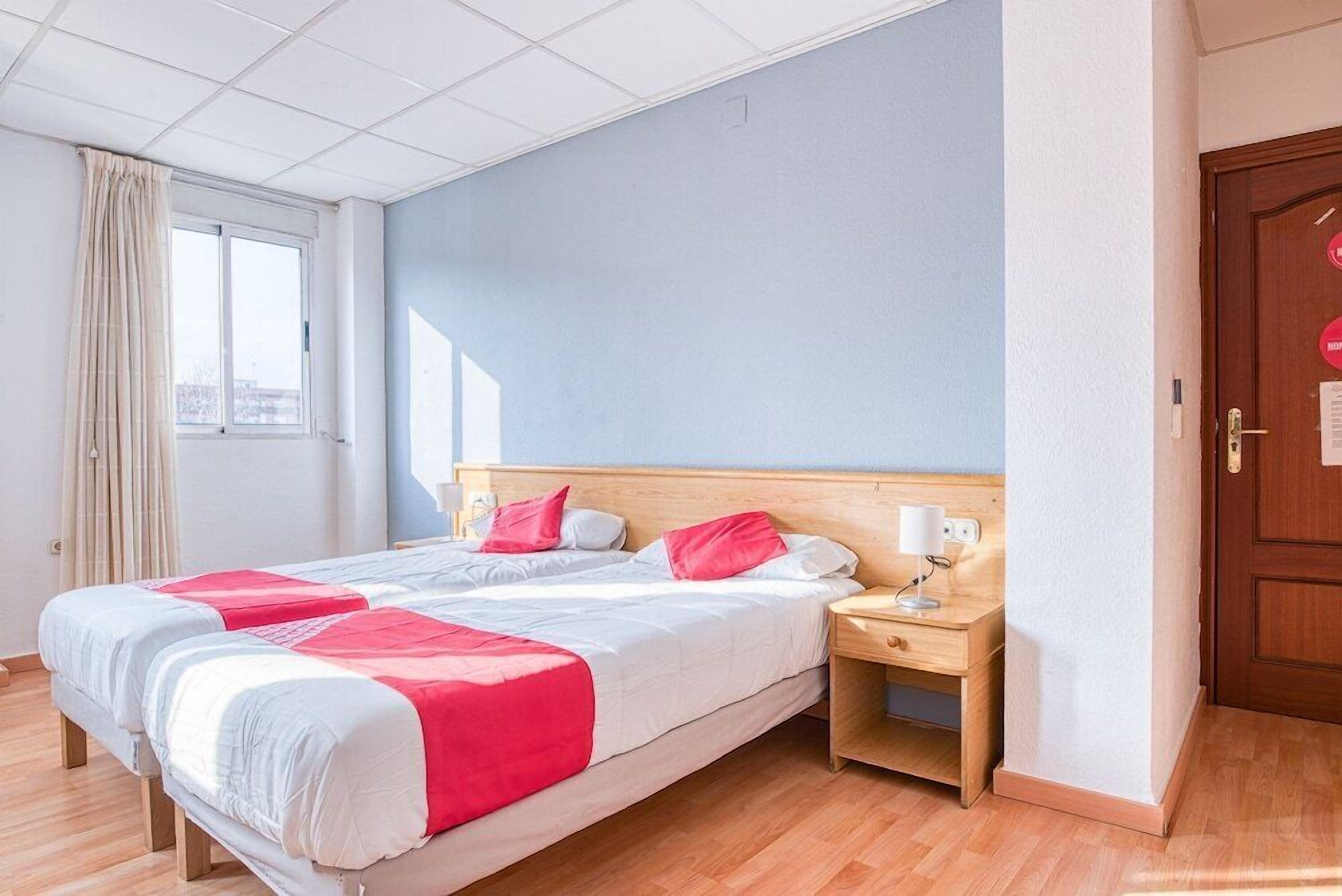 Una habitación de hotel con una cama grande, dos mesitas de noche, una ventana con cortinas y una puerta de madera.