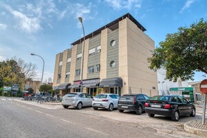 Un hotel de varios pisos con un diseño moderno, que cuenta con una planta baja con restaurante y un hostal. Varios coches están estacionados al frente, y se observan árboles y un cielo despejado al fondo.