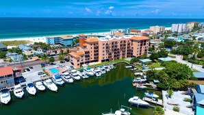A marina with multiple boats docked, a hotel with balconies overlooking the marina, and a beach in the background.