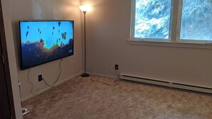 A room with a flat-screen TV mounted on the wall, a standing floor lamp, a window with a view of snow-covered trees, and a beige carpet. - Wonderfully located two bedroom apartment (Pierre)
