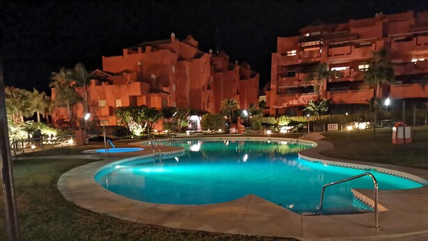 Ein Schwimmbecken, umgeben von einer gut beleuchteten Fläche, im Hintergrund ein Gebäude.