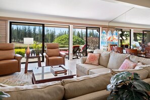A spacious living room with a sofa, armchairs, and a coffee table. Large windows offer a view of a garden and trees. - New Listing: Spacious 2-bedroom Luxurious Kapalua Golf Villa (Lahaina)
