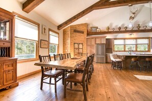 Un comedor espacioso con una larga mesa de madera y sillas, una cocina con electrodomésticos modernos y una pared rústica de piedra con la inscripción "The Gathering Place".