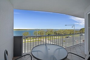 Un balcon avec une table et des chaises, donnant sur une étendue d’eau et une route où circulent des véhicules.