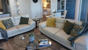 Un salon moderne avec deux canapés en cuir blanc, une table basse en verre et une petite table d’appoint sur laquelle est posée une plante. Une grande fenêtre laisse entrer la lumière naturelle, et un ventilateur de plafond est visible.
