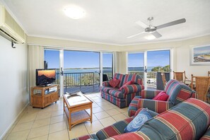 A spacious living room with a large sofa set, a television, and a ceiling fan. - Sandy Shores Luxury Holiday Units (Sunshine Coast)