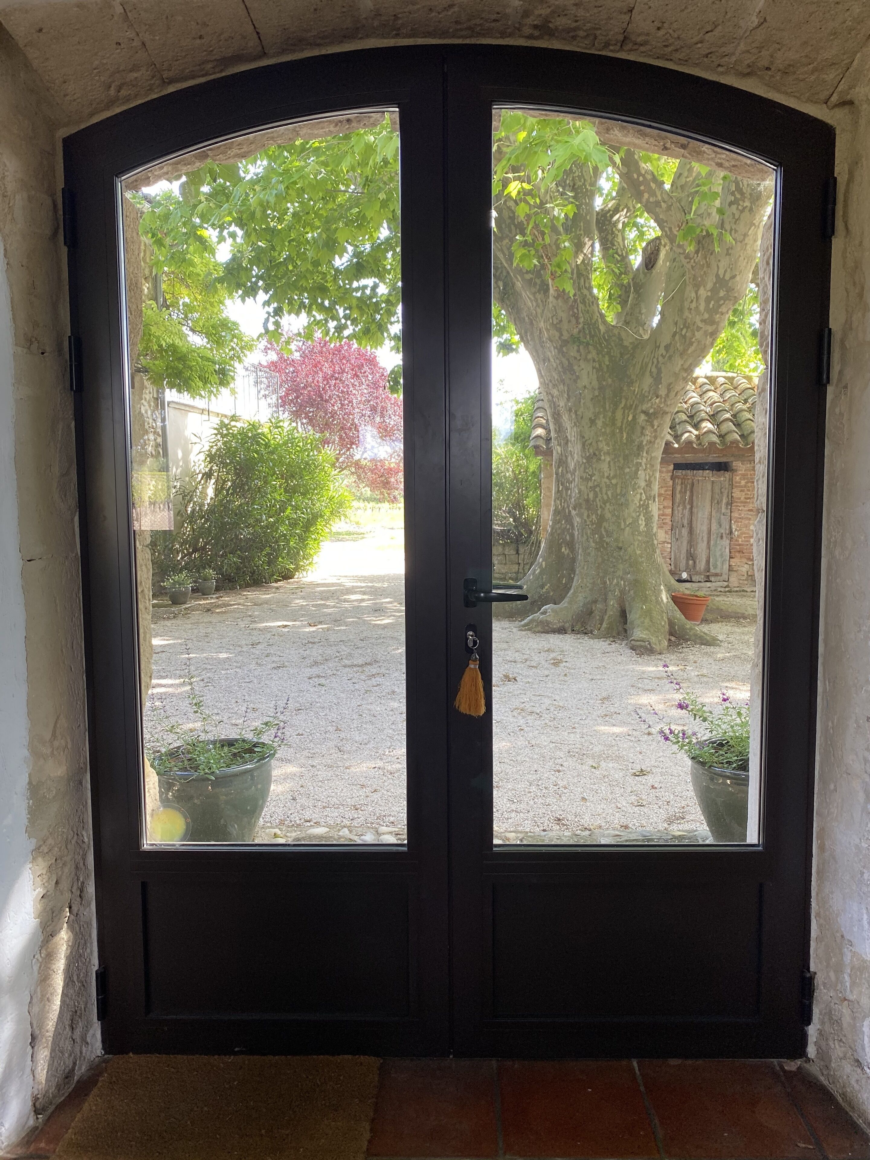 Une porte en bois sombre, dotée de panneaux de verre, qui donne sur un espace recouvert de gravier, agrémenté de plantes en pot et d’un arbre.