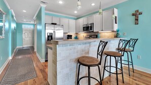 A kitchen with white cabinets, a stainless steel refrigerator, a microwave, and a bar area with stools. - The Wharf Resort 423 | Food-Shopping-Entertainment | My Beach Getaways (Orange Beach)