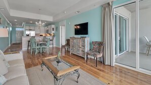 A spacious living room with a sofa, a coffee table, and patterned chairs. A television is mounted on the wall, and there is a sliding glass door leading to a balcony. - The Wharf Resort 423 | Food-Shopping-Entertainment | My Beach Getaways (Orange Beach)