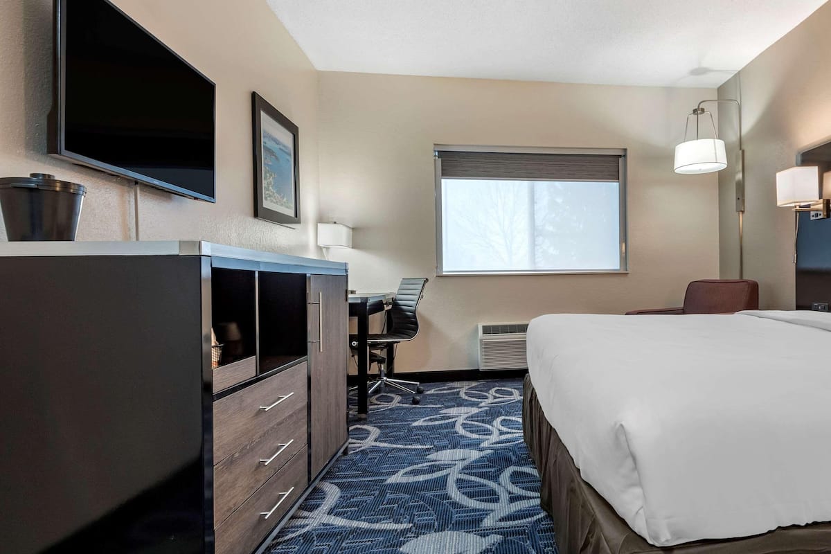 A hotel room with a bed, a desk with a chair, a television, a window with blinds, and a lamp.