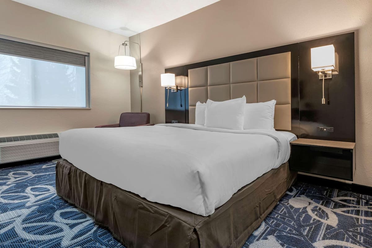 A hotel room with a large bed, a bedside table, a chair, a window with blinds, and a wall-mounted light fixture.